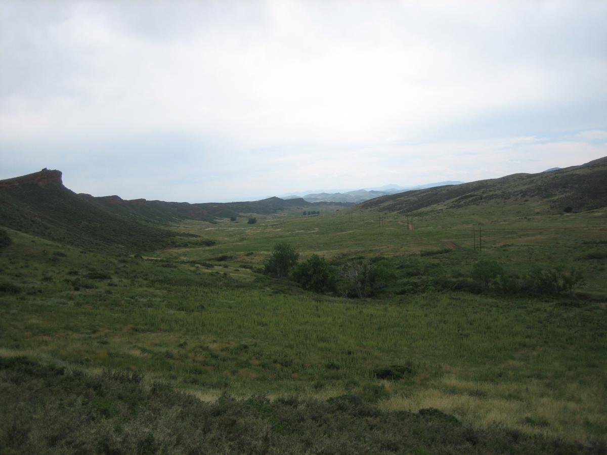 A panoramic view of a green valley bordered by rolling hills under a cloudy sky, with some distant mountains visible in the background. The landscape features a mix of grass and shrubs, and a few trees can be seen scattered throughout the area. Devil's Backbone mountain bike trail.
