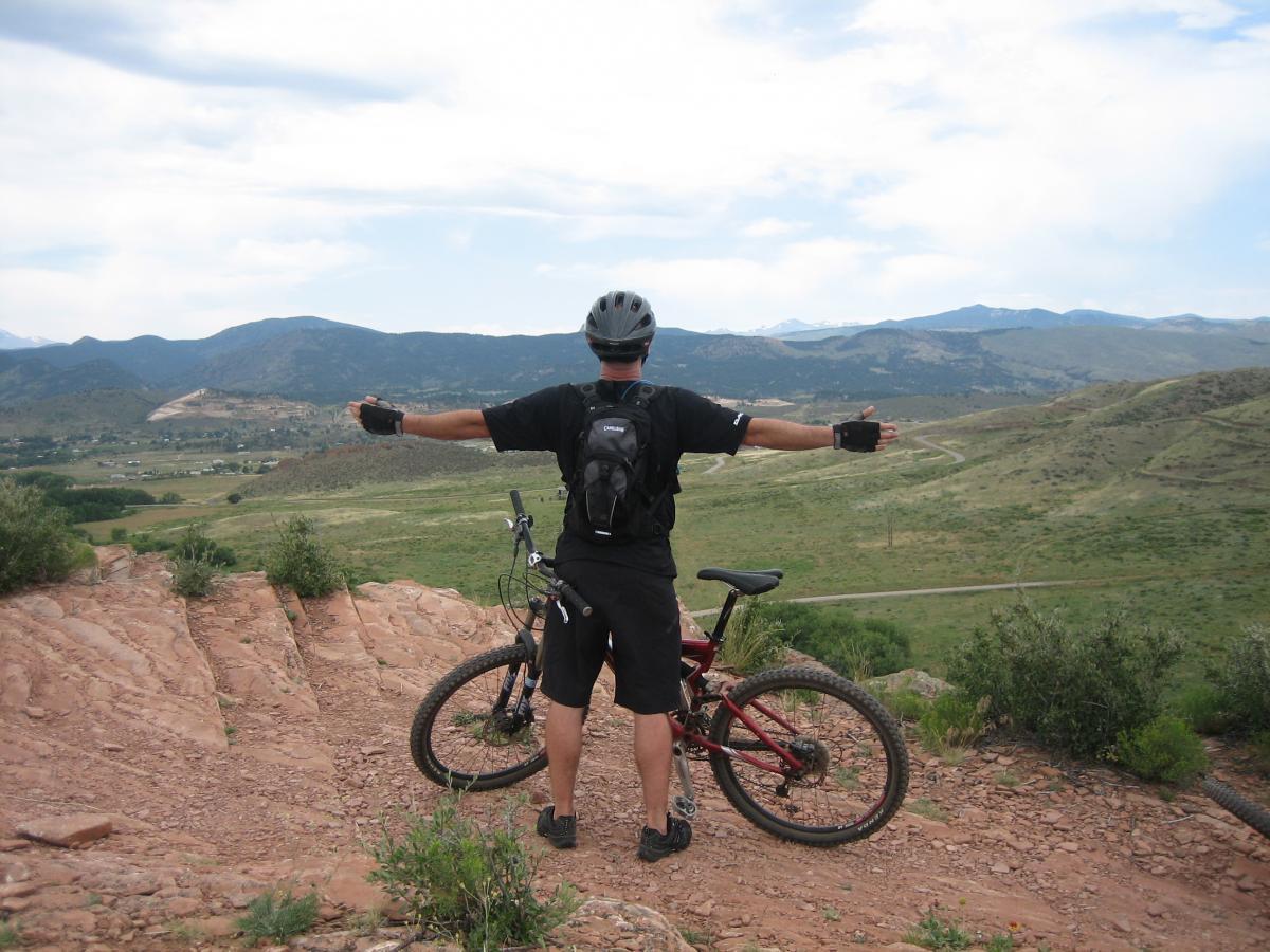 A mountain biker stands with arms outstretched on a rocky ledge, overlooking a vast landscape of rolling hills and distant mountains under a cloudy sky. The bike rests beside him, and the scene captures a sense of freedom and adventure in nature. Devil's Backbone mountain bike trail.