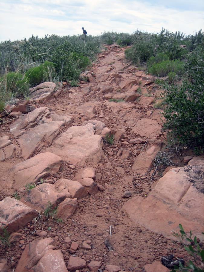 A rocky hiking trail winding through green shrubs and grass, leading up to a distant figure at the top of a hill under a cloudy sky. Devil's Backbone mountain bike trail.