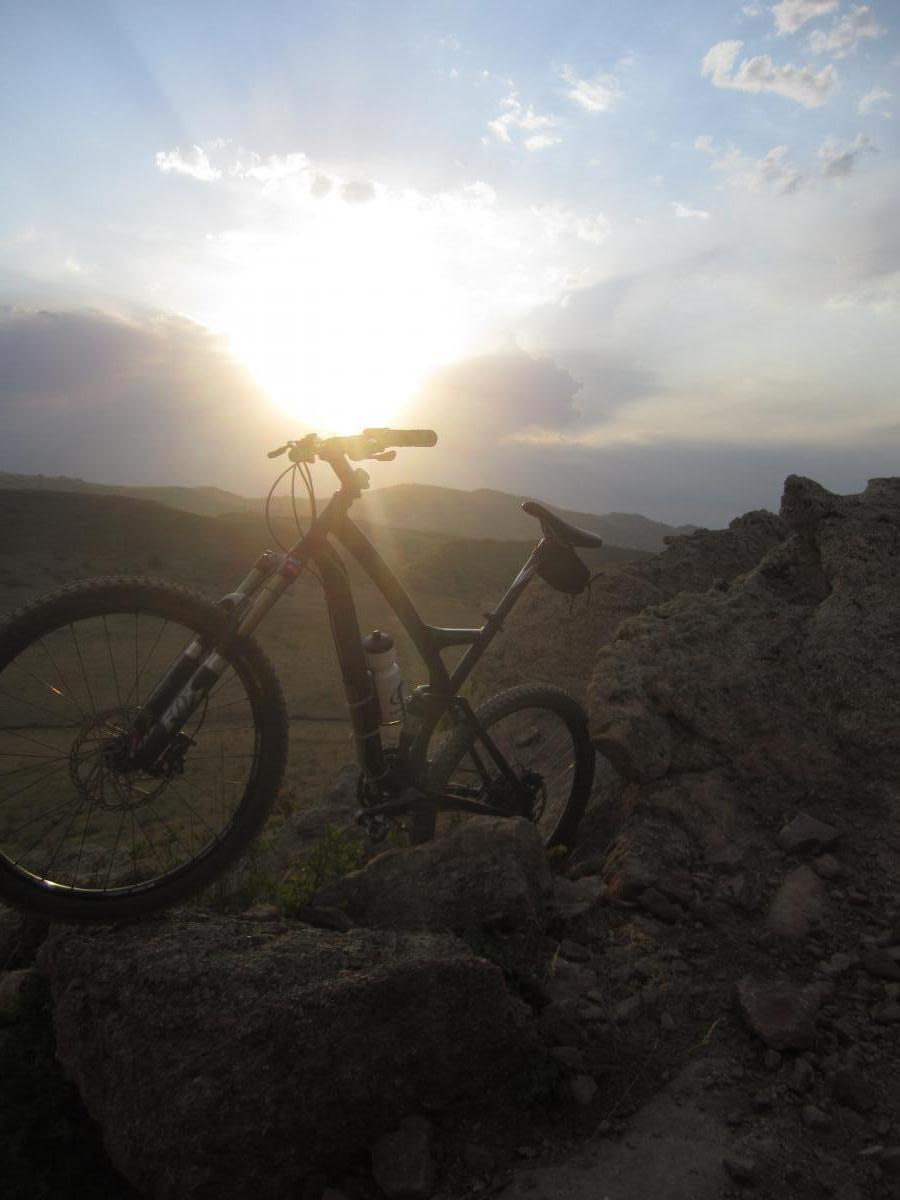 A mountain bike resting on rocks with a sunset in the background, casting a warm glow over the landscape. The skies are partially cloudy, and the sun is positioned low, creating dramatic lighting across the scene. Coyote Ridge mountain bike trail.
