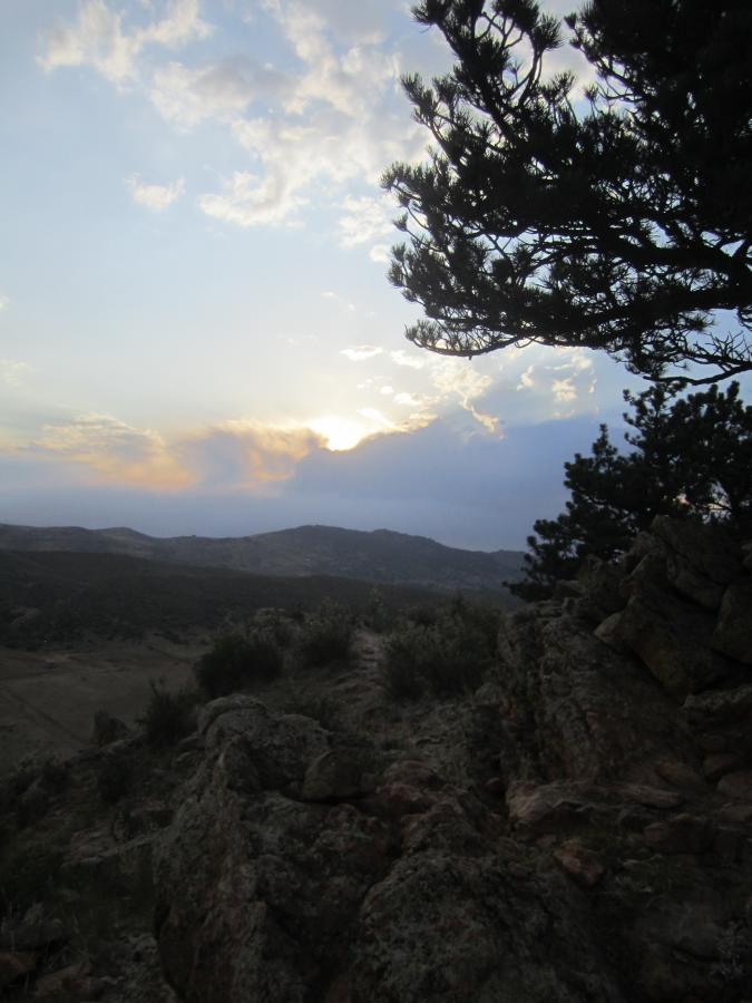 A scenic view of a mountainous landscape during sunset, featuring a cloudy sky with shades of orange and blue. In the foreground, a silhouette of a pine tree and rocky terrain contrasts with the bright horizon. Coyote Ridge mountain bike trail.