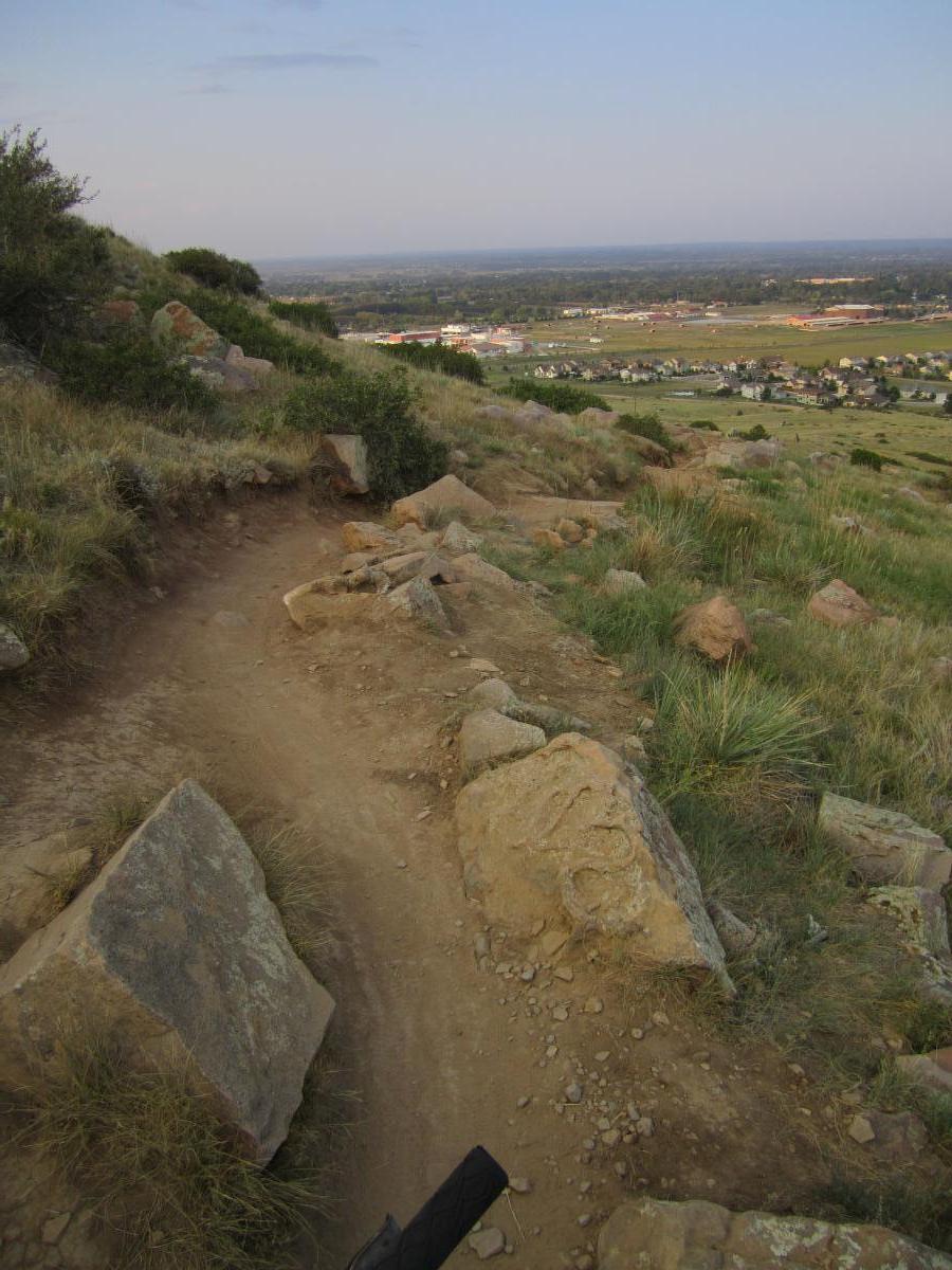 A scenic view from a hillside trail, featuring a dirt path winding through rocky terrain and sparse grass. In the background, a panoramic view reveals a valley with buildings and green fields under a clear sky, suggesting a tranquil outdoor setting. Maxwell's mountain bike trail.