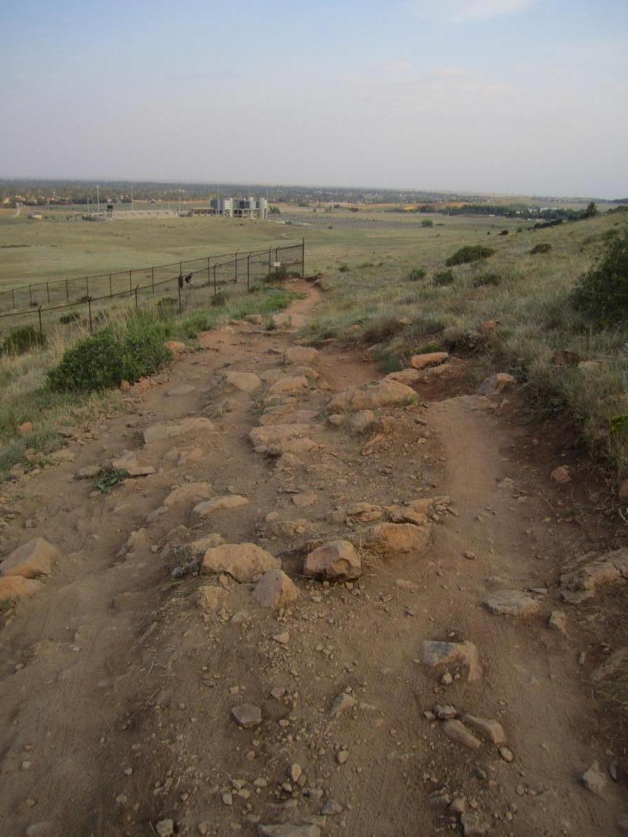 A dirt path surrounded by rocky terrain leads down a hill, with a view of a distant structure and open fields under a cloudy sky. Fencing is visible along the path, suggesting the border of an area with natural vegetation and sparse grass. Maxwell's mountain bike trail.