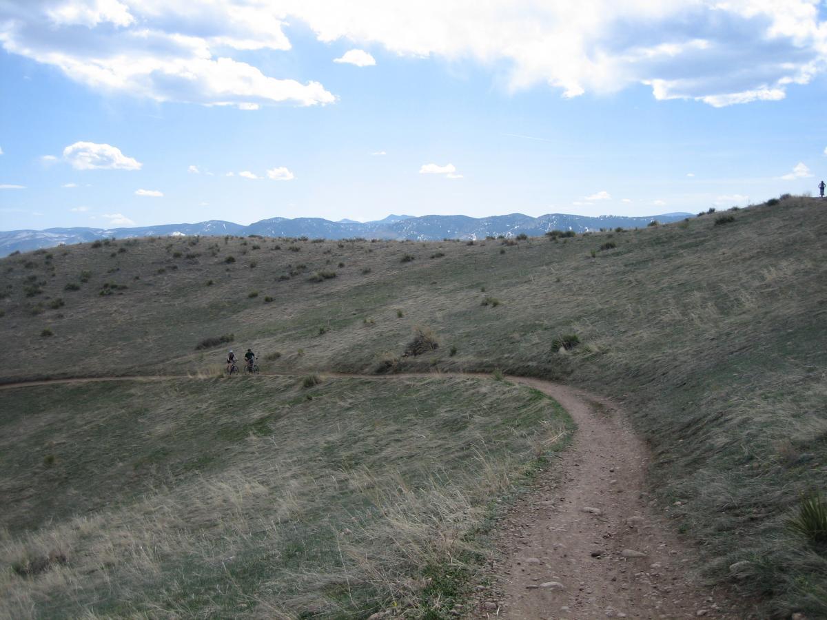 A winding dirt path leads through a grassy hillside under a blue sky with scattered clouds. Two people on bicycles ride along the trail, while a distant figure stands on a nearby rise, with mountains visible in the background. Flatirons Vista mountain bike trail.