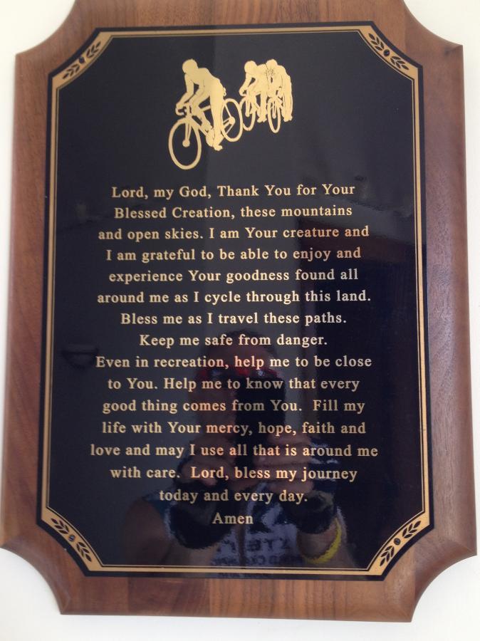 A wooden plaque with a gold inscription featuring a prayer expressing gratitude for nature and a request for safety and guidance while cycling. The design includes the silhouettes of cyclists at the top, and the text emphasizes appreciation for creation, the importance of recreation, and a plea for blessings during journeys. Kingdom Trails mountain bike trail.