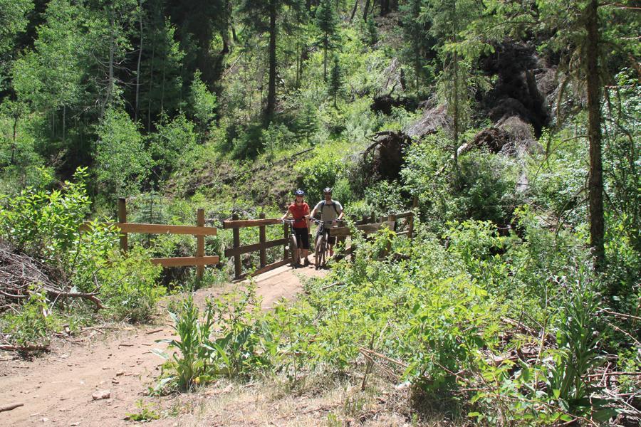 Two mountain bikers stand at a wooden bridge in a lush, green forest. The scene features dense vegetation, including trees and bushes, typical of a natural outdoor trail. The bikers are wearing helmets and casual outdoor clothing, appearing to take a break while enjoying the surrounding nature. The path is dirt, leading through the vibrant foliage. Hermosa Creek Trail mountain bike trail.