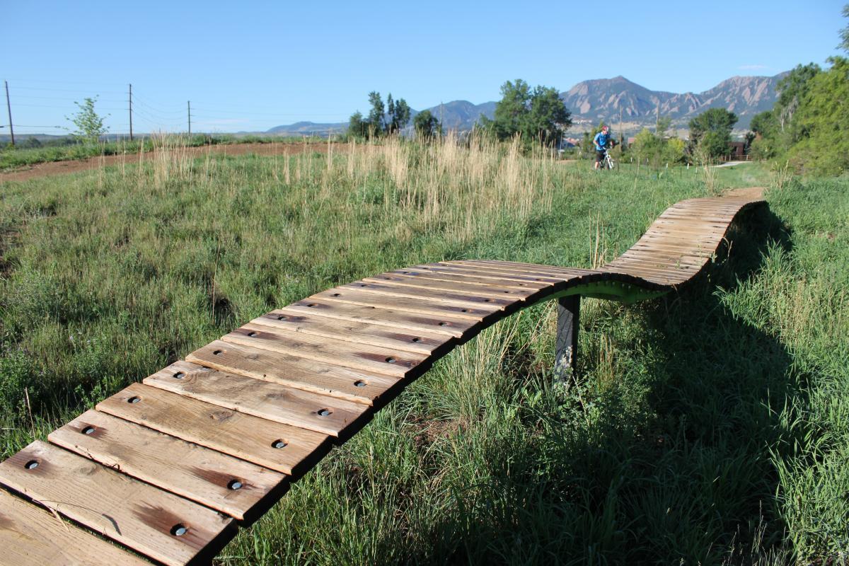 A curving wooden plank bridge elevated above grassy terrain, surrounded by tall grass and scattered shrubs, with mountains visible in the background under a clear blue sky. A person on a bicycle can be seen in the distance, riding along a path. Valmont Bike Park mountain bike trail.