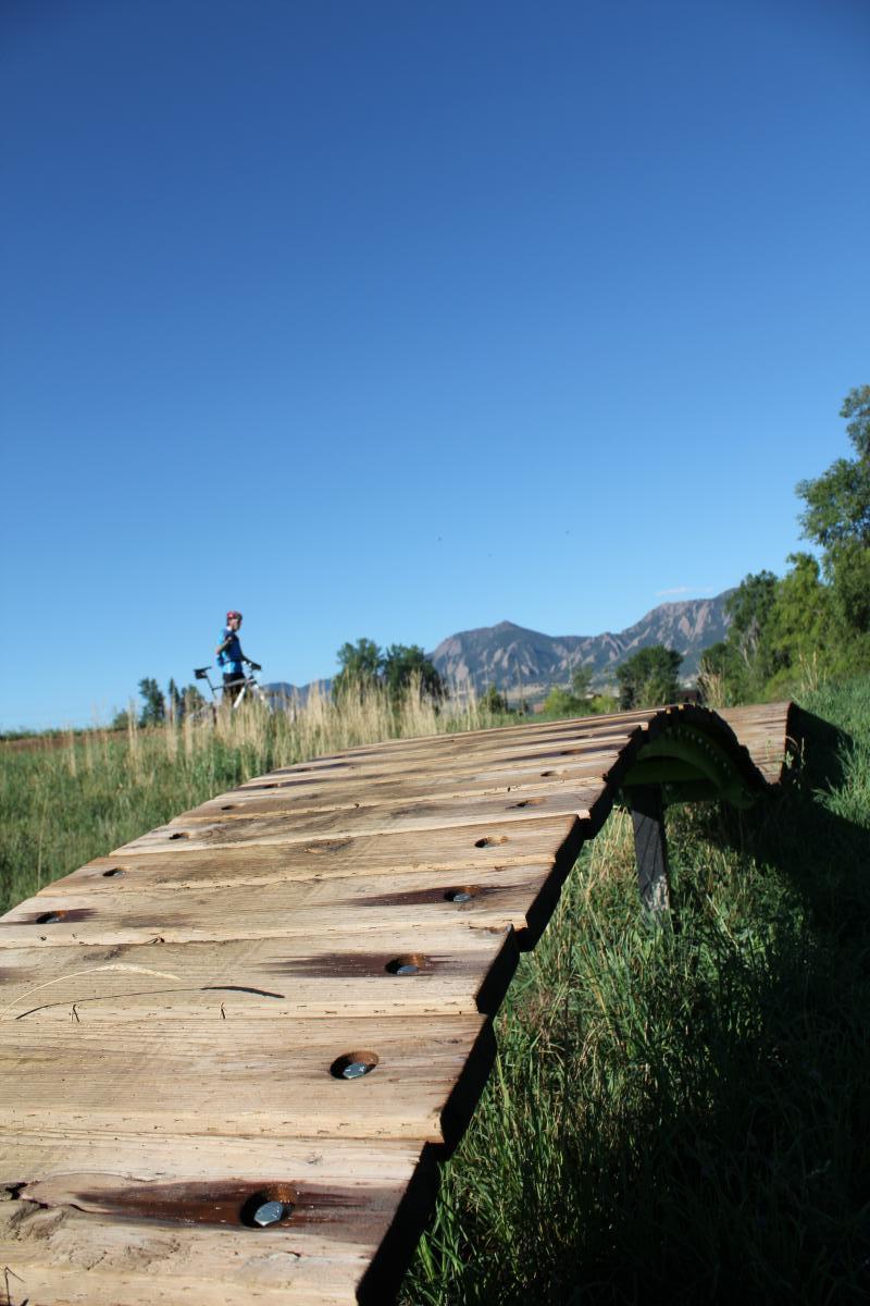 A wooden bike ramp curves through tall grass, with a clear blue sky overhead and mountains in the background. A person is seen riding a bike in the distance, adding a sense of activity to the outdoor scene. Valmont Bike Park mountain bike trail.