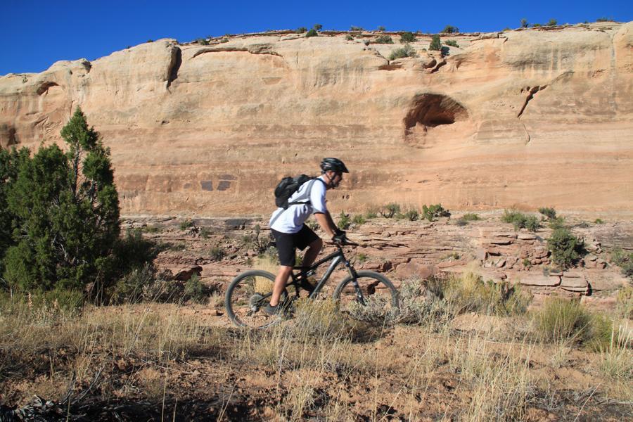 A mountain biker rides along a rocky trail in front of a large sandstone cliff, with sparse vegetation and blue sky in the background. Mary's Loop / Horsethief Bench mountain bike trail.