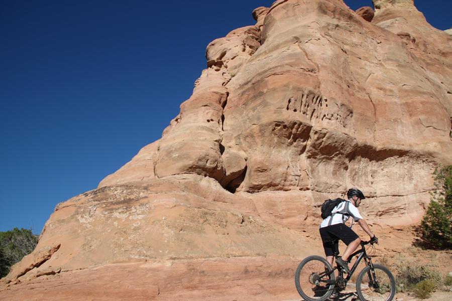 A mountain biker rides along a dirt trail next to a large, reddish rock formation under a clear blue sky. The scene captures the rugged terrain and natural beauty of the landscape, highlighting the adventurous spirit of outdoor cycling. Mary's Loop / Horsethief Bench mountain bike trail.