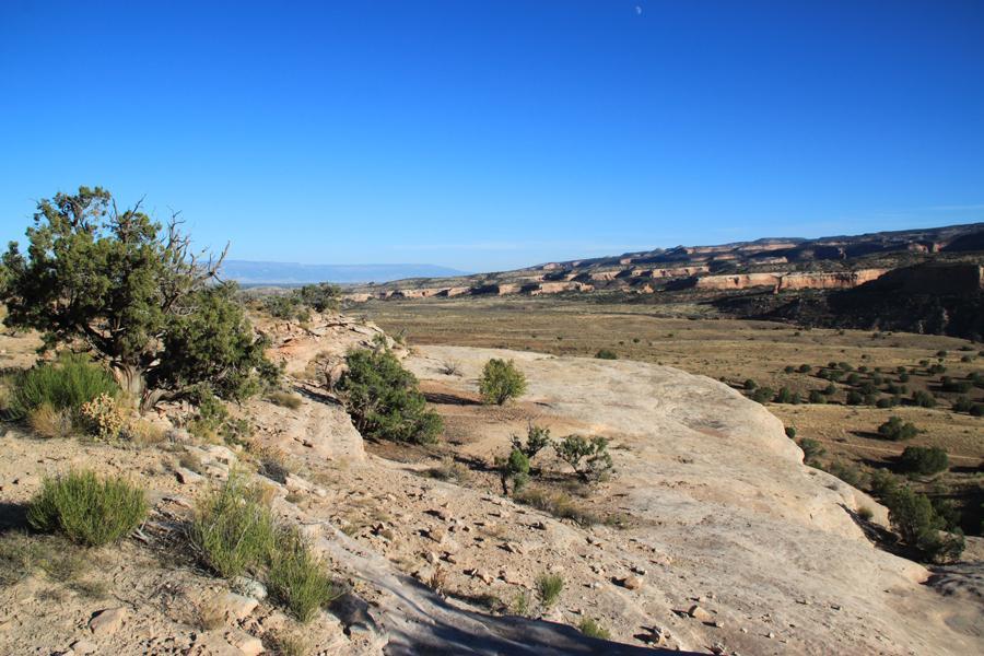 A rugged landscape featuring a rocky outcrop and sparse vegetation, with a clear blue sky above. Rolling hills and distant cliffs are visible in the background, suggesting a vast and arid terrain. Mary's Loop / Horsethief Bench mountain bike trail.