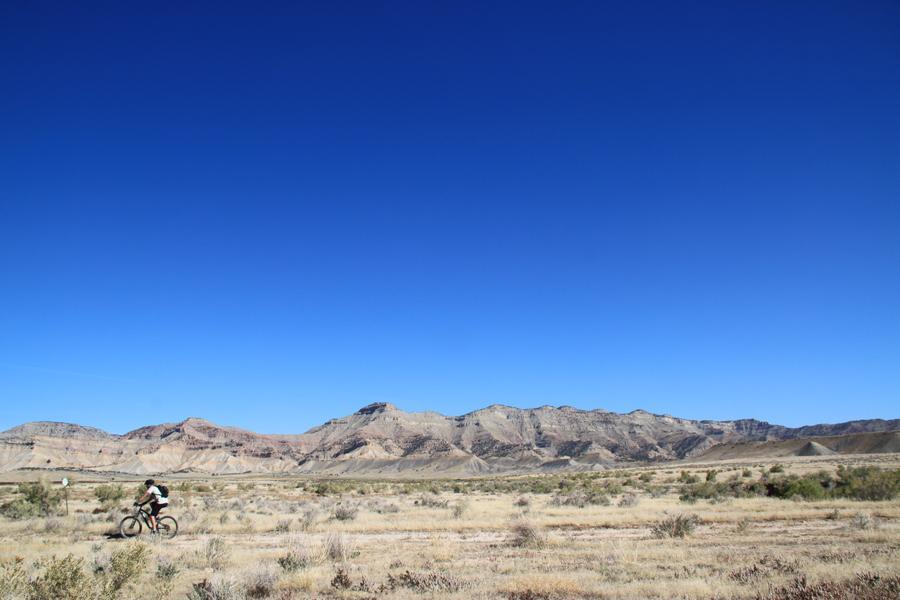 A mountain biker riding on a dirt path in a vast, arid landscape with layered brown mountains in the background under a clear blue sky. Zippety Do Dah mountain bike trail.