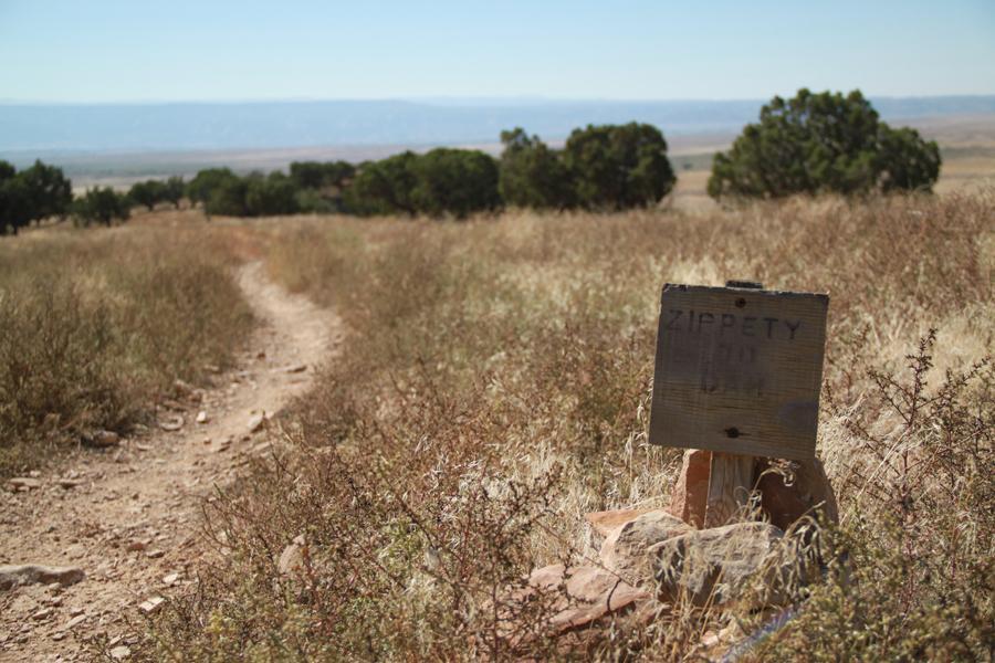 A dirt trail winding through tall grass and shrubs, marked by a weathered wooden sign that reads "Zippety" in a rustic font. In the background, rolling hills and a clear blue sky stretch into the distance. Zippety Do Dah mountain bike trail.
