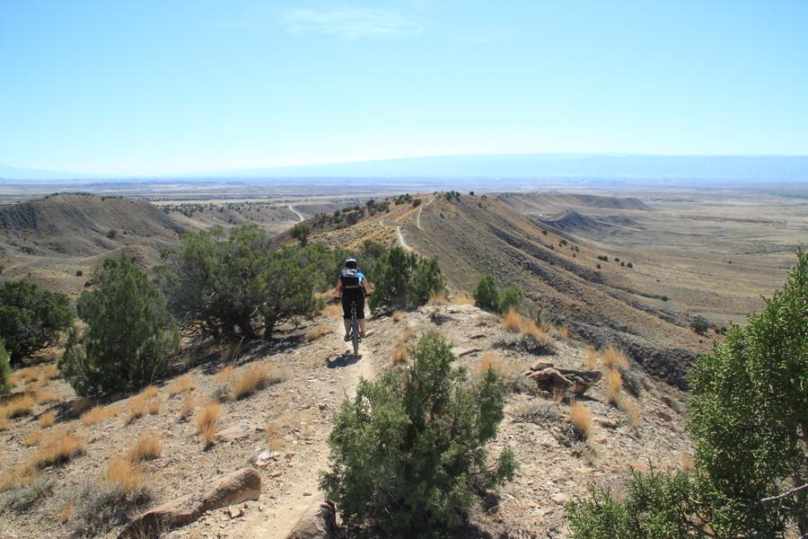 A person riding a mountain bike on a trail along a ridge, surrounded by dry, rugged terrain and sparse vegetation. The landscape features rolling hills and a clear blue sky, showcasing an expansive view of the valley below. Zippety Do Dah mountain bike trail.