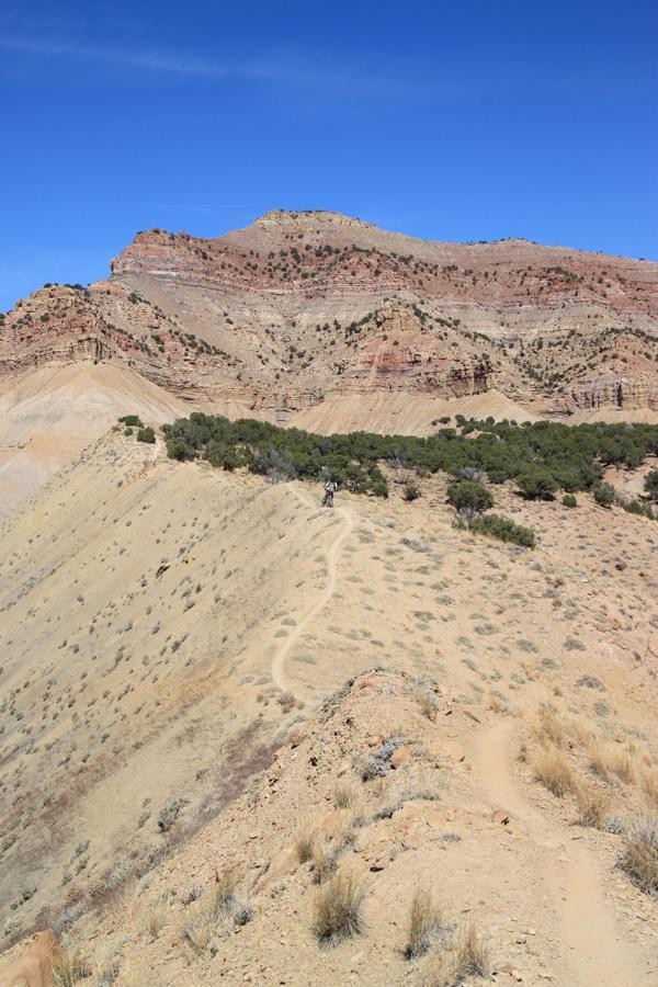 A winding dirt trail meanders through a rugged, arid landscape, flanked by a backdrop of layered, colorful rock formations under a clear blue sky. A lone hiker is visible along the trail, surrounded by sparse vegetation and slopes of tan and brown earth. Zippety Do Dah mountain bike trail.