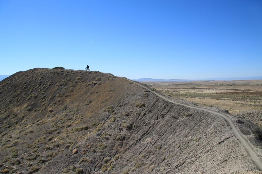 A lone cyclist rides on a dirt path at the top of a rocky hill, surrounded by wide open plains and a clear blue sky. The landscape features rolling hills and sparse vegetation, emphasizing the tranquility of the outdoor setting. Zippety Do Dah mountain bike trail.