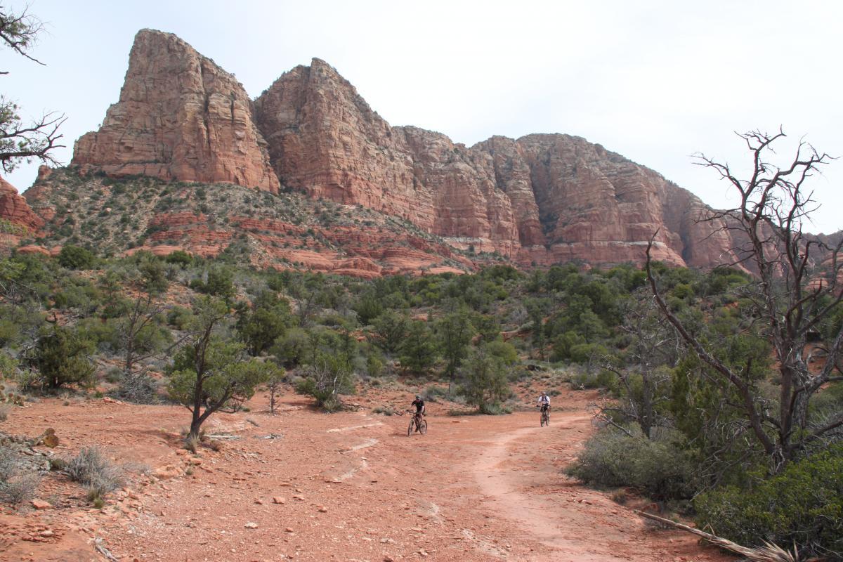 A scenic view of a rocky landscape with towering red cliffs in the background. Two mountain bikers traverse a dirt path that winds through a sparsely wooded area, featuring shrubs and scrubby trees against a rugged terrain. The sky is overcast, adding a serene atmosphere to the outdoor scene. Broken Arrow Trail / Chicken Point mountain bike trail.
