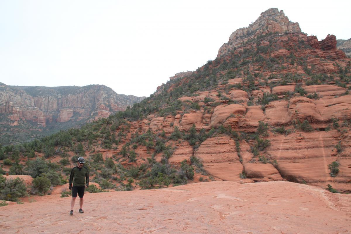 A person standing on a reddish rocky terrain with hills and green shrubs in the background. The sky is overcast, and towering red rock formations rise in the distance. The individual is wearing a helmet and casual outdoor clothing, looking down at the ground. Broken Arrow Trail / Chicken Point mountain bike trail.