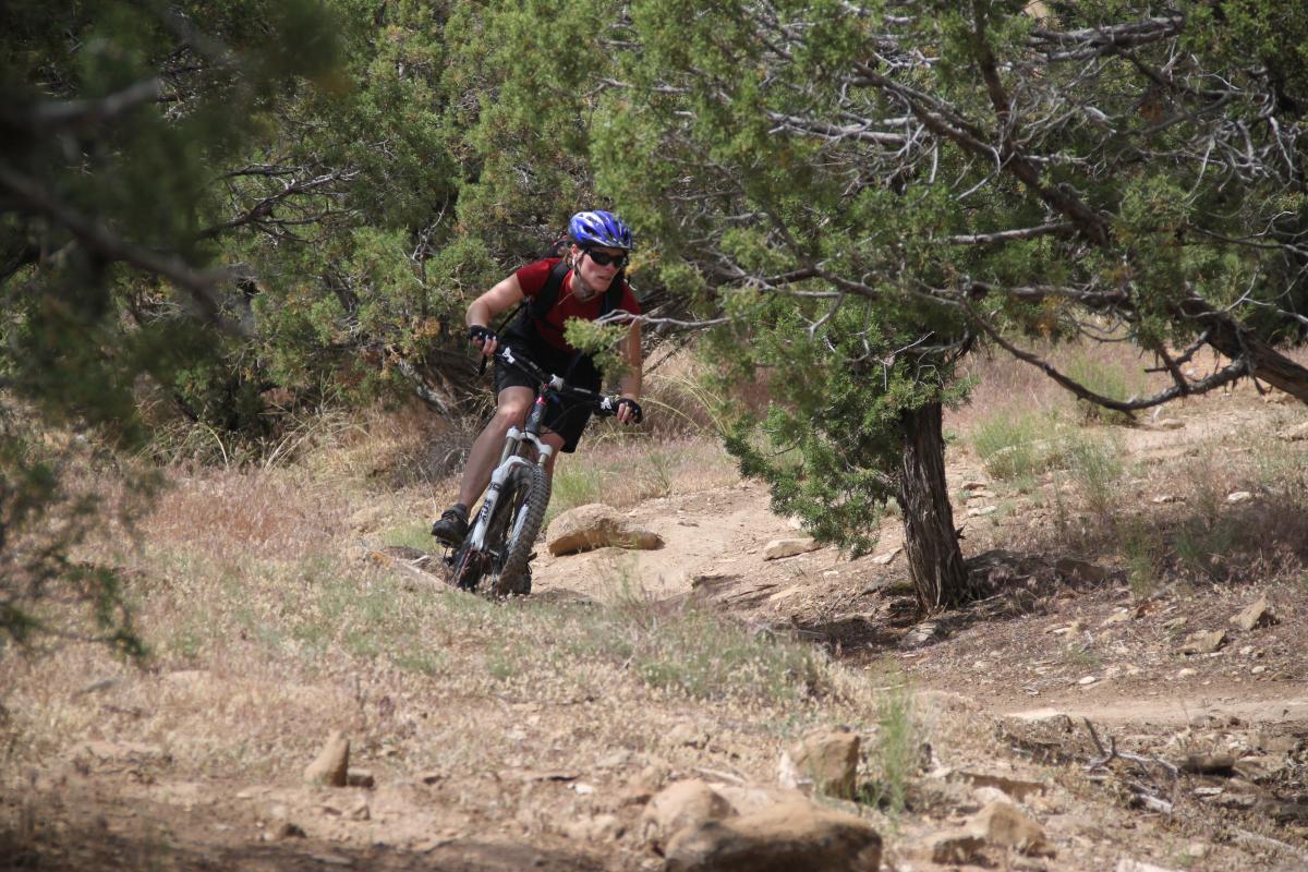 A mountain biker navigating a rocky trail surrounded by trees, wearing a helmet and sunglasses, and focused on the path ahead. Kessel Run mountain bike trail.