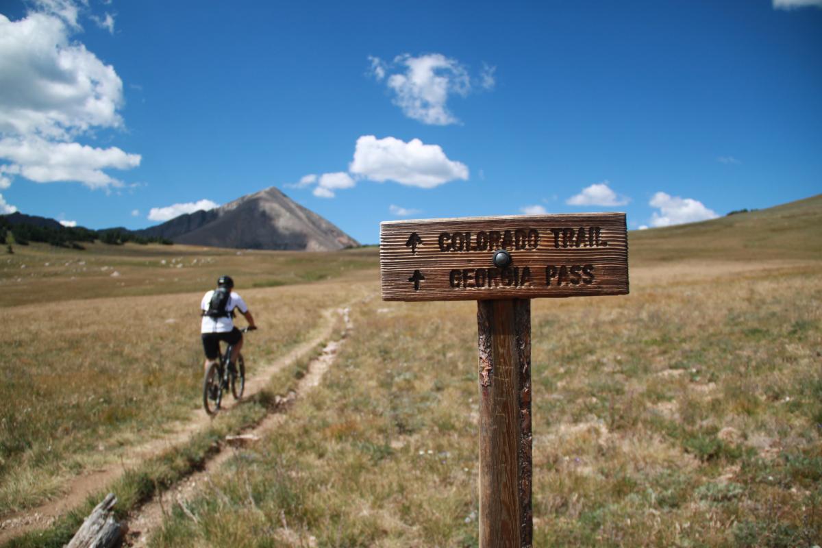 A mountain biker riding on a dirt trail through a grassy area, with a wooden trail sign indicating directions to "Colorado Trail" and "Georgia Pass." In the background is a mountain under a blue sky with scattered clouds. Georgia Pass 4x4 Road mountain bike trail.