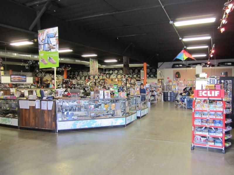 Interior of an outdoor gear shop featuring glass display cases filled with various products. The space has dark ceilings and is well-lit, with shelves lined with bicycles, equipment, and merchandise. Posters and banners are visible on the walls, and there