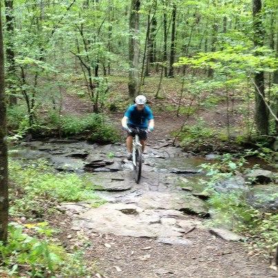 A person riding a mountain bike over rocky terrain in a lush green forest. Nockamixon State Park mountain bike trail.
