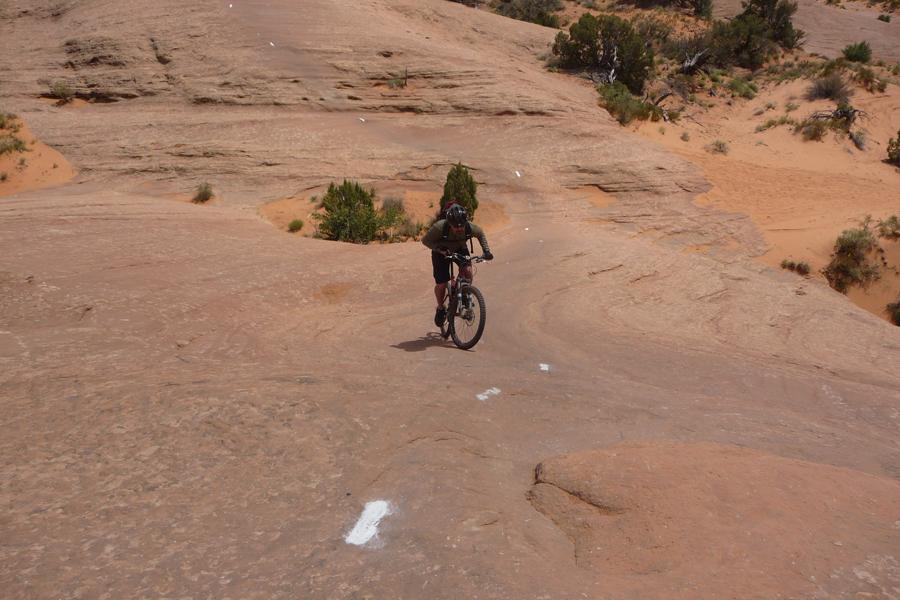 A person riding a mountain bike on a rocky terrain, with a sandy area in the background. The rider is wearing a helmet and casual athletic clothing, navigating a sloped surface marked by white directional indicators. Vegetation, including small bushes and trees, is visible nearby. Slickrock mountain bike trail.