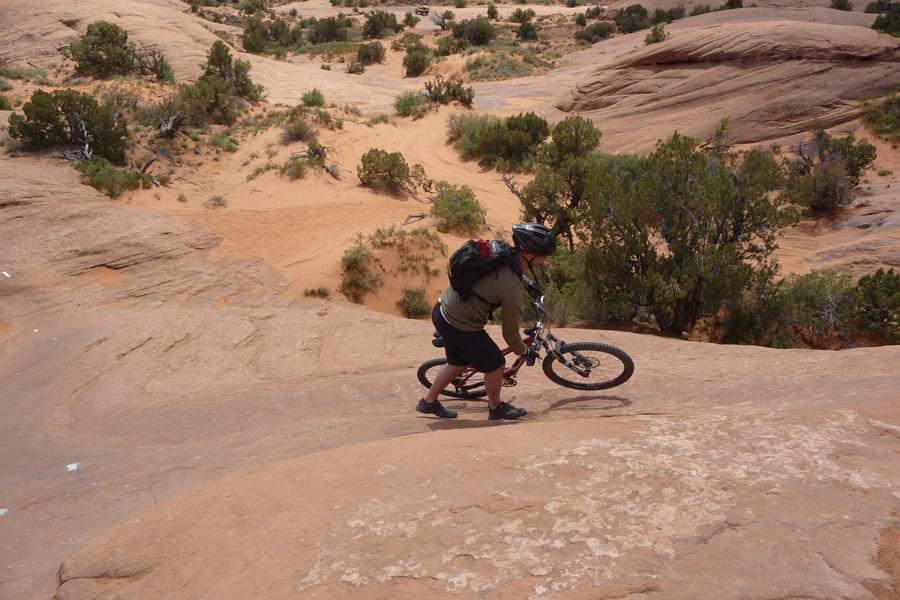 A person in outdoor gear pushing a mountain bike uphill on a rocky terrain, surrounded by desert vegetation and sandy areas. Slickrock mountain bike trail.