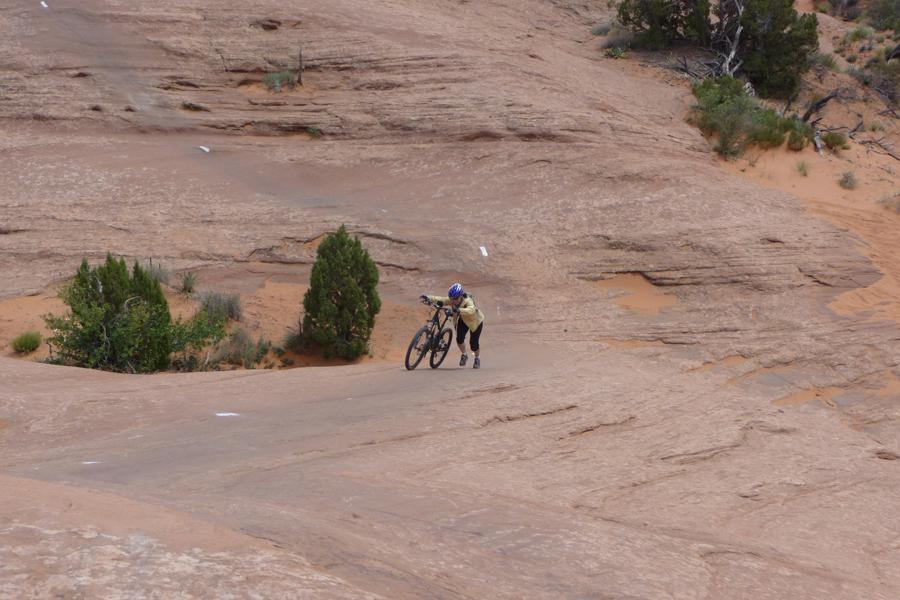 A person wearing a helmet and cycling gear is struggling to push a mountain bike up a steep, rocky slope in a desert landscape. Sparse vegetation is visible in the background, along with orange sand and rugged rock formations. Slickrock mountain bike trail.