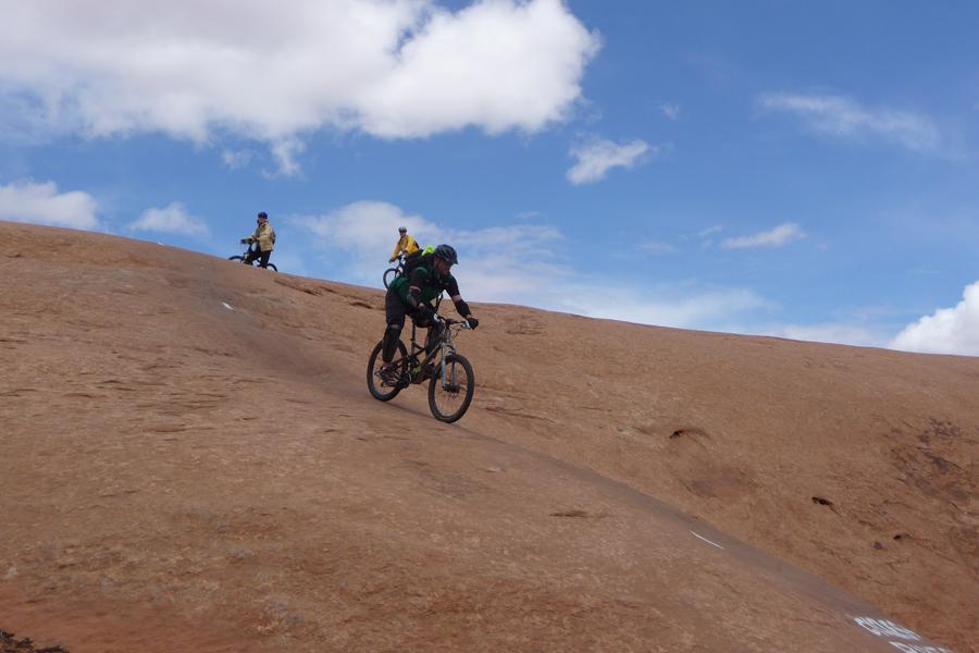 A mountain biker descending a rocky terrain with red earth under a blue sky, while two other cyclists are visible in the background. Slickrock mountain bike trail.