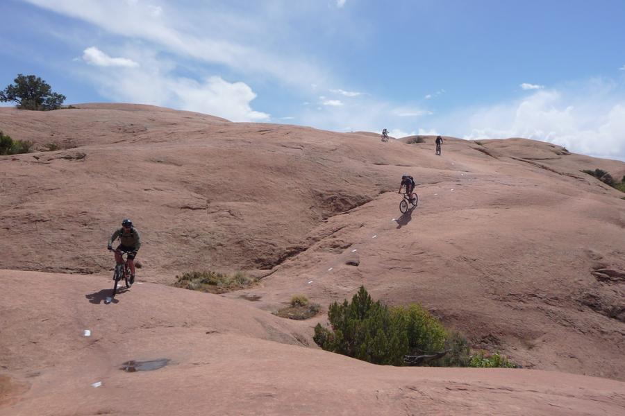 Bikers navigating rocky terrain under a blue sky with white clouds in a desert landscape. The path features white markers on the reddish rock surface, showcasing an adventurous outdoor setting. Slickrock mountain bike trail.