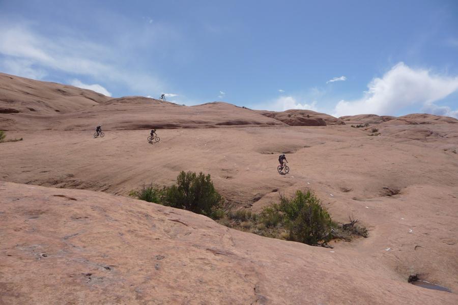 Three mountain bikers traverse a rocky, arid landscape under a blue sky with scattered clouds. The terrain is characterized by smooth, reddish-brown rock formations and sparse vegetation. Slickrock mountain bike trail.