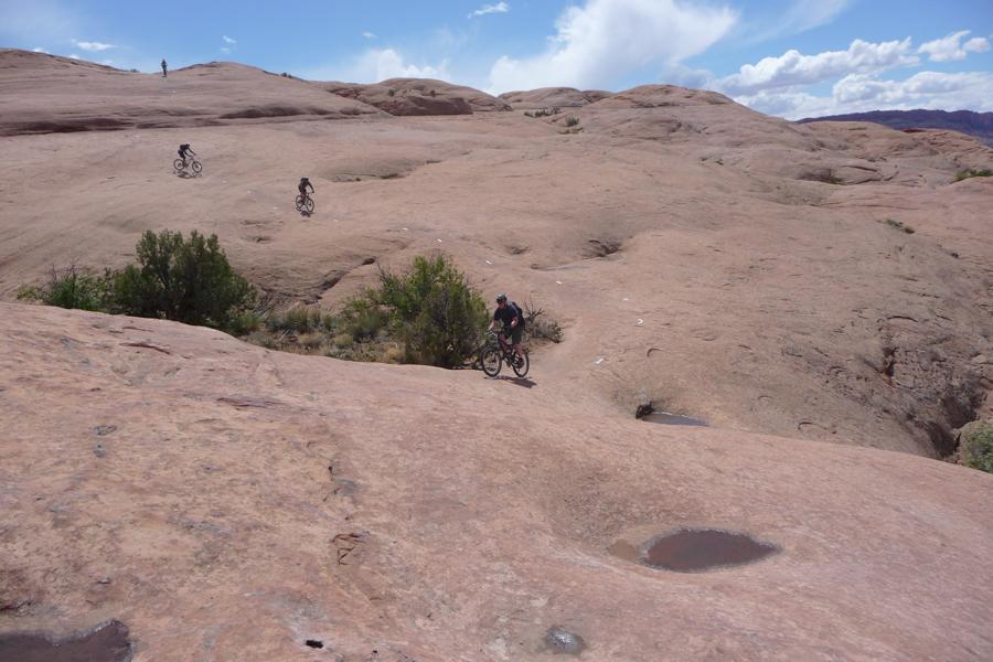 Alt text: Four mountain bikers are riding over a rocky, desert-like terrain with sparse vegetation under a blue sky with clouds. Slickrock mountain bike trail.