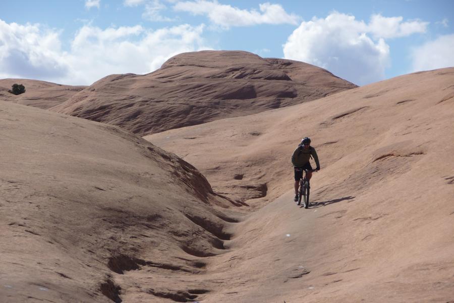 A person riding a mountain bike along a rocky path in a desert landscape, surrounded by smooth, sandstone-like formations under a partly cloudy sky. Slickrock mountain bike trail.