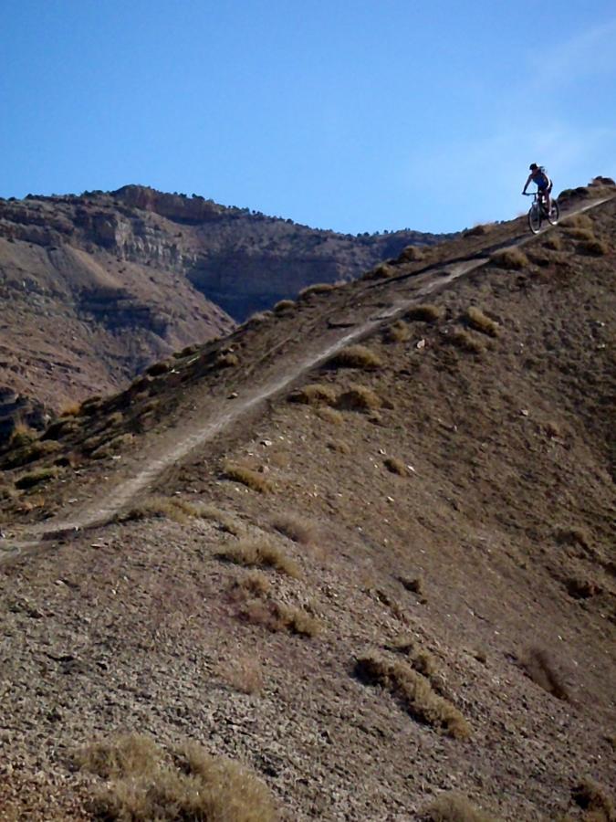 A mountain biker riding along a narrow dirt trail on a steep hillside, with rocky terrain and a clear blue sky above. The backdrop features rugged mountains and sparse vegetation. 18 Road Trails / North Fruita Desert mountain bike trail.