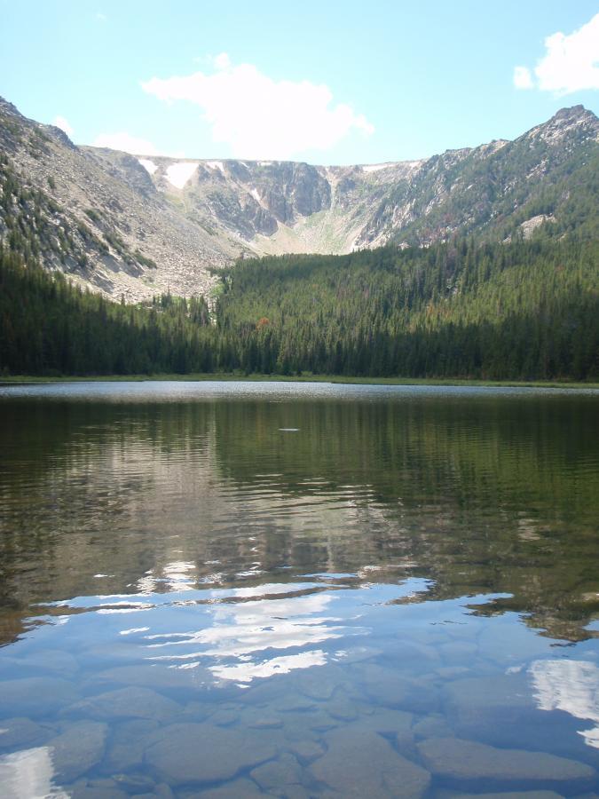 A serene mountain lake surrounded by dense green forests and rocky cliffs under a blue sky with fluffy white clouds. The clear water reflects the mountains, creating a peaceful and scenic landscape. Silver And Basin Lakes Trail mountain bike trail.