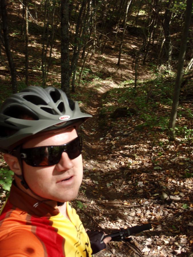 A person wearing a helmet and sunglasses takes a selfie while mountain biking on a wooded trail. The background features trees with autumn leaves scattered on the ground, hinting at a sunny day in nature. Standing Rocks mountain bike trail.