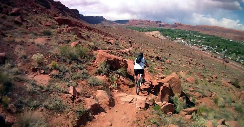A mountain biker navigates a rocky and rugged trail in a desert landscape, with distant hills and a small town visible in the valley below under a partly cloudy sky. Pipe Dream mountain bike trail.