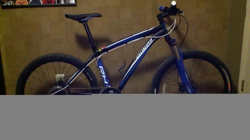 Specialized Rockhopper Comp Disc: A blue mountain bike with large tires is propped against a wall. The bike features a sleek frame design, front suspension, and a mounted light on the handlebars. The background shows textured wallpaper, adding to the indoor setting.