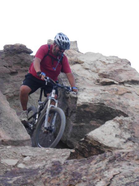 A person in a red shirt and helmet is riding a mountain bike down a rocky, uneven trail. The landscape features large boulders and rugged terrain, indicating an adventurous outdoor setting. Lunch Loops mountain bike trail.
