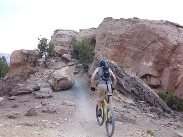 A person riding a mountain bike on a rocky trail surrounded by large boulders and sparse shrubbery. The cyclist is wearing a helmet and a backpack, navigating a steep ascent in a rugged outdoor environment. Lunch Loops mountain bike trail.