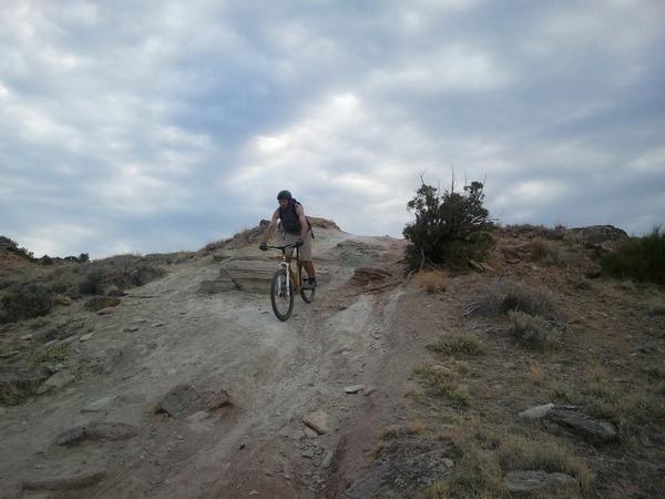 A person riding a mountain bike on a rocky, sloped terrain under a cloudy sky. The landscape features sparse vegetation and boulders, indicating an outdoor adventure setting. Lunch Loops mountain bike trail.