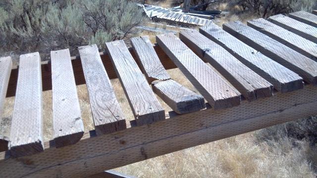 Wooden walkway with missing and weathered planks, surrounded by dry grass and shrubbery. The structure appears to be in need of repair. Ada/Eagle Bike Park mountain bike trail.
