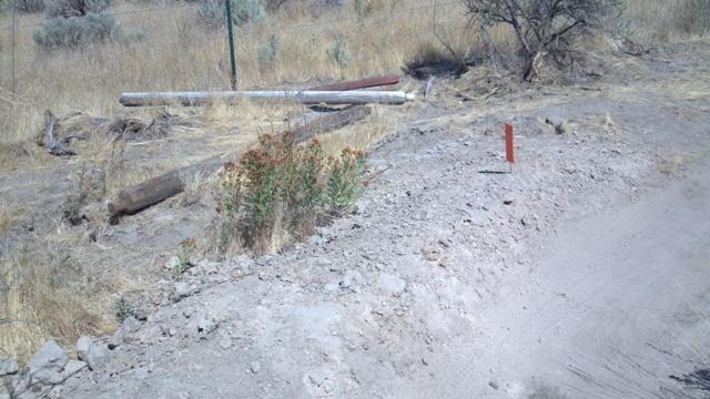 A dry, barren landscape with sparse vegetation, featuring a dirt path on the right. In the foreground, there are patches of greenery and a small red marker. In the background, a few wooden poles lie on the ground alongside dry grass and shrubs. Ada/Eagle Bike Park mountain bike trail.