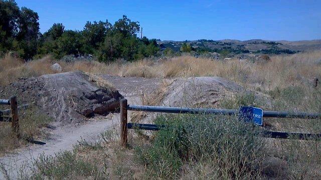 A dirt pathway leading into a grassy, overgrown field, flanked by two dirt mounds and a wooden fence with a sign indicating access. In the background, rolling hills and trees are visible under a clear blue sky. Ada/Eagle Bike Park mountain bike trail.