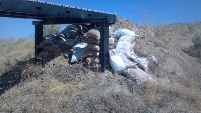 A pile of sandbags leaning against a small dirt mound, located beneath a black, constructed platform in a dry, open landscape with sparse vegetation. Ada/Eagle Bike Park mountain bike trail.