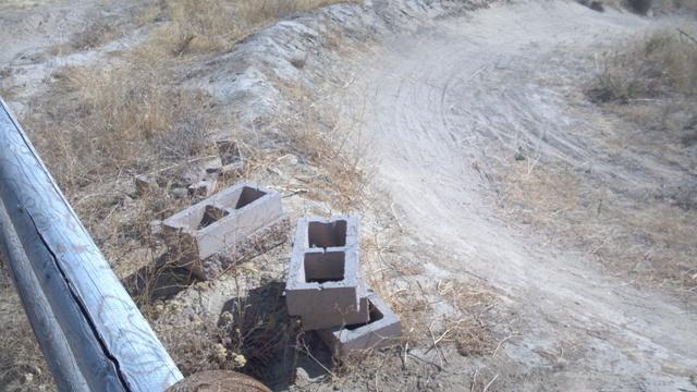 Two concrete blocks are located on a dry, dusty path with sparse dry grass on either side. A metal railing is partially visible on the left side of the image. The path curves in the background, suggesting a rural or abandoned area. Ada/Eagle Bike Park mountain bike trail.