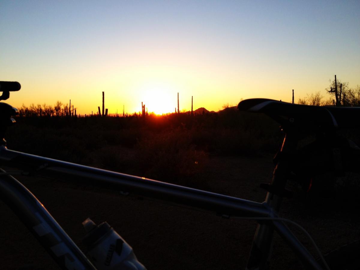 A silhouette of a bicycle against a vibrant sunset, with cacti and desert plants in the background. The sun is setting on the horizon, casting warm golden and orange hues across the sky. Pima Road and Dynamite Blvd mountain bike trail.