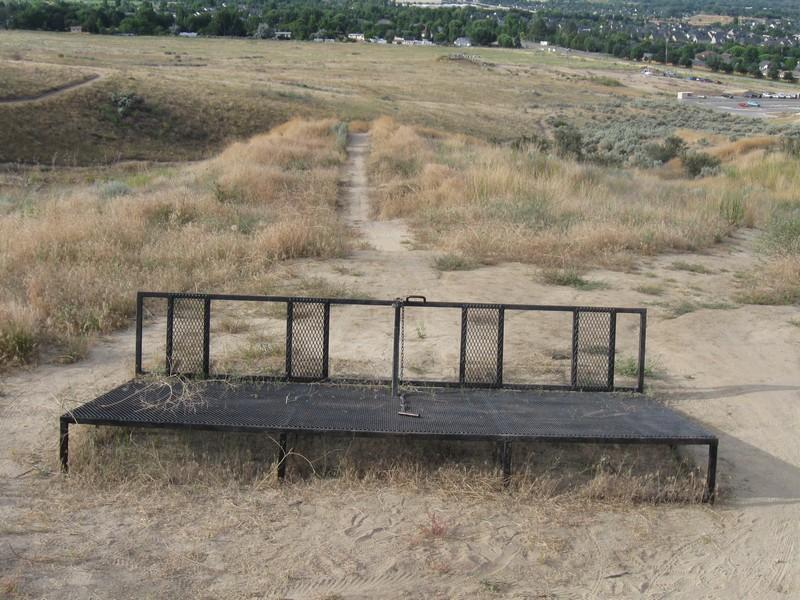A metal platform surrounded by dry grass and dirt, situated on a hillside with a winding trail leading down. In the background, there are patches of trees and a distant view of buildings and hills. The scene captures an outdoor area that appears to be partly neglected or overgrown. Ada/Eagle Bike Park mountain bike trail.