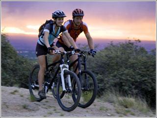 Two mountain bikers ride side by side on a trail at sunset, surrounded by greenery. The sky displays vibrant hues of orange and purple. Both riders wear helmets and cycling gear, appearing engaged and enjoying their ride.